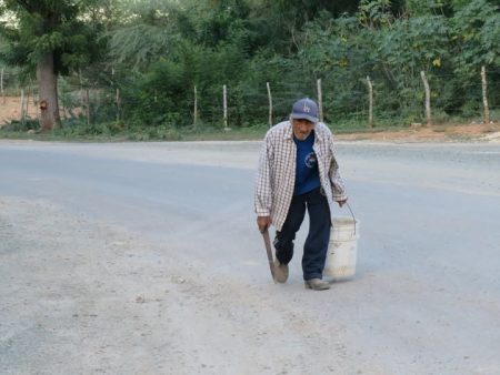 En Manzanillo: Consejo de Organizaciones lanza campaña solicitando respaldo nacional e internacional para que Mecanismo de Investigación del BID declare elegible reclamo del caso Manzanillo Bay Energy Texto alternativo: Pescador de edad avanzada camina rumbo a Estero Balsa, por una carretera de Manzanillo cargando un cubo y una herramienta de pesca, símbolo de las dificultades actuales para acceder a las zonas tradicionales de pesca.